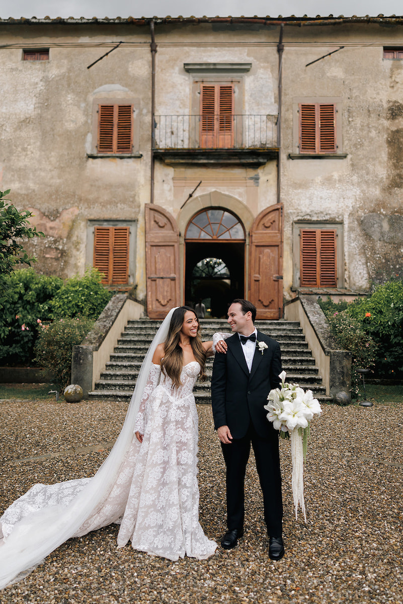Bride and groom stand smiling in front of an old building with open wooden doors, dressed in wedding attire, holding a bouquet and a glass.