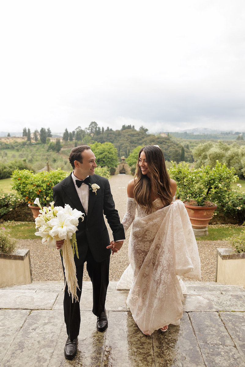 A couple in formal attire walks hand in hand outdoors, with greenery and a scenic landscape in the background. The man holds a bouquet of white flowers.