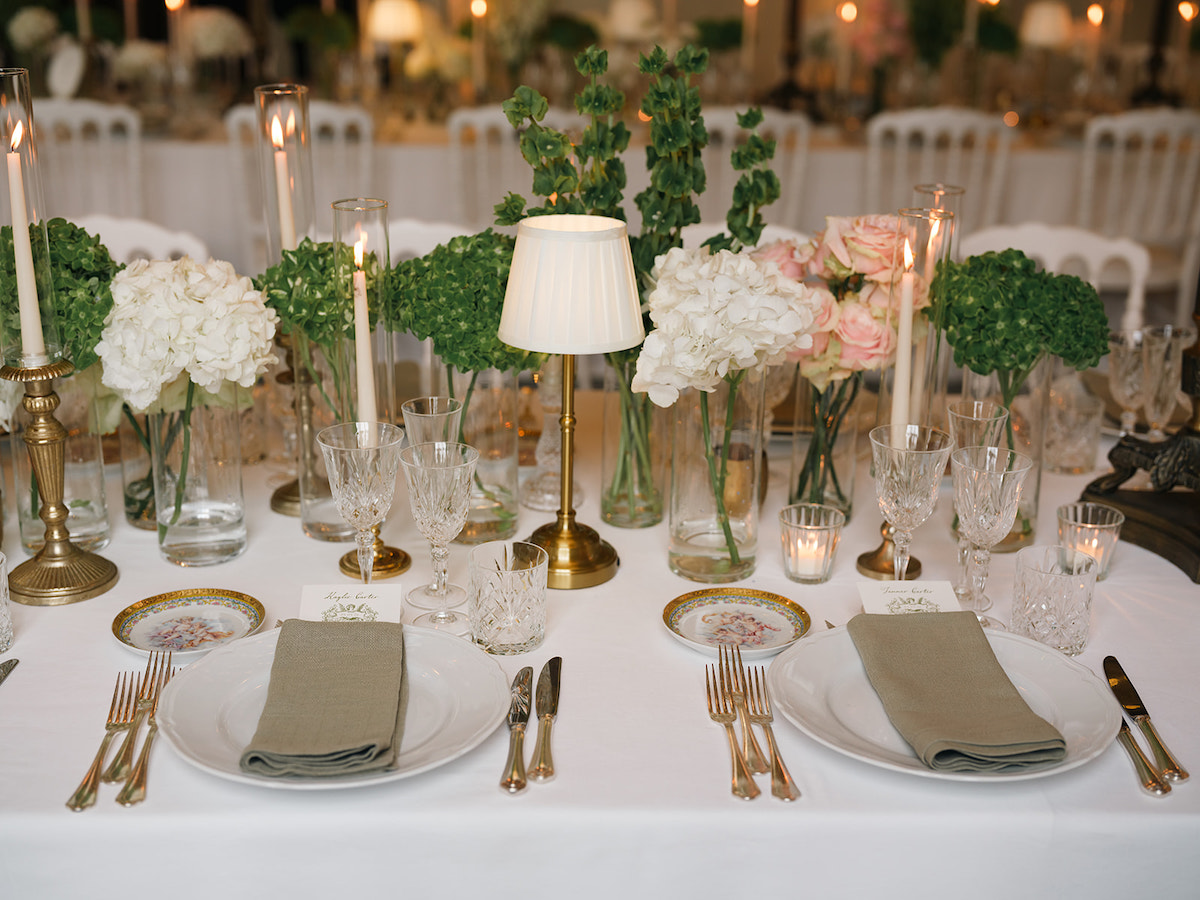 Elegant table setting with white plates, beige napkins, cutlery, glassware, candlesticks, a small lamp, and vases of white and green flowers on a white tablecloth.