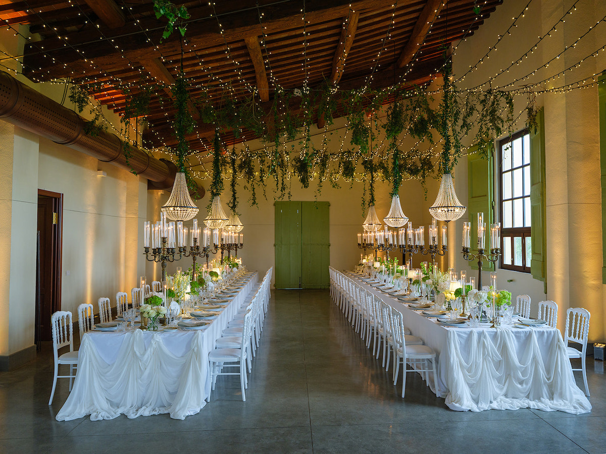 Two long banquet tables with white tablecloths, elegant chandeliers, candles, and greenery are arranged in a spacious, decorated hall with high ceilings and hanging plants.