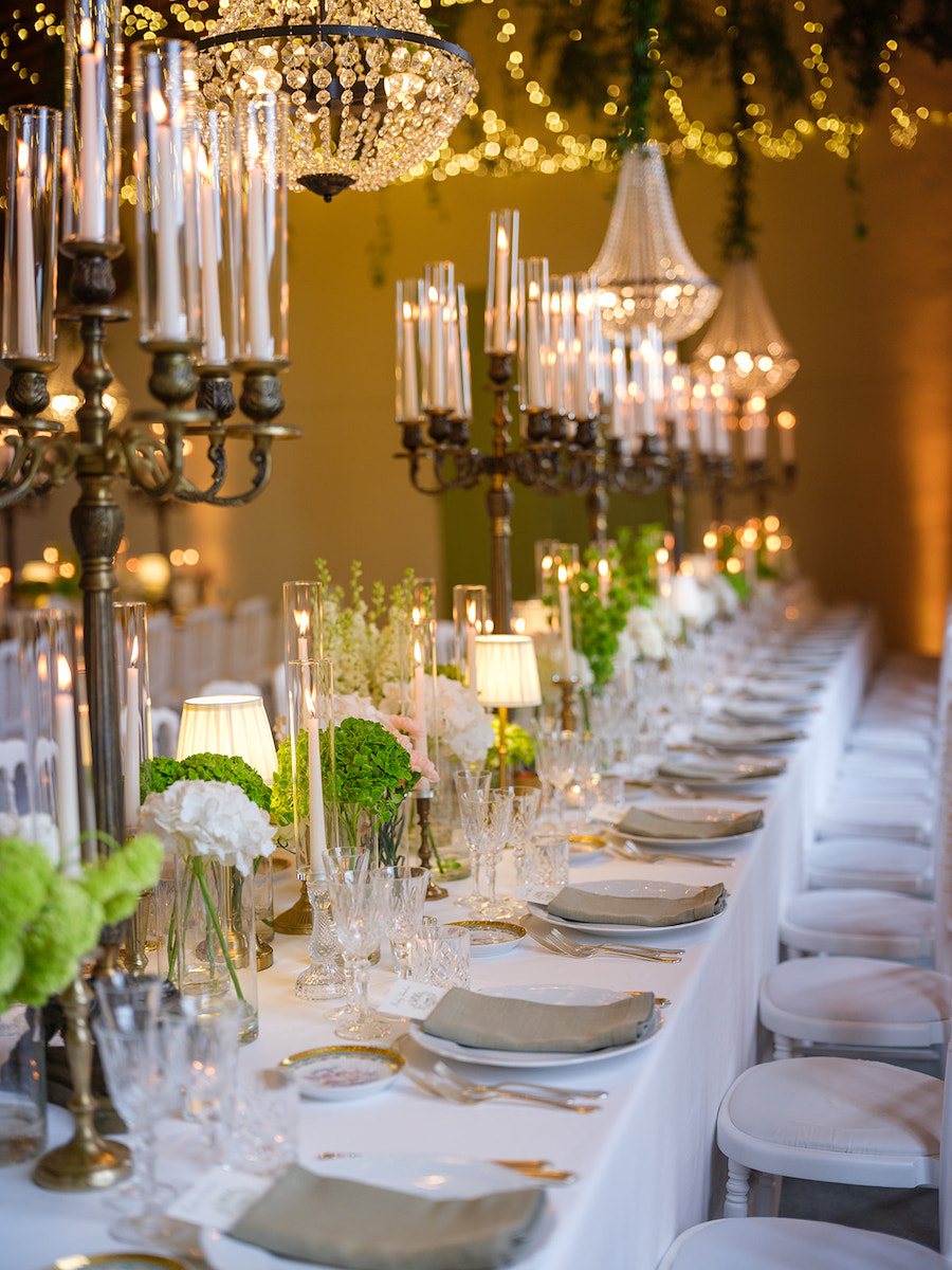 Elegant banquet table set with white linens, plates, glassware, green floral arrangements, and tall candelabras, under chandeliers and string lights.