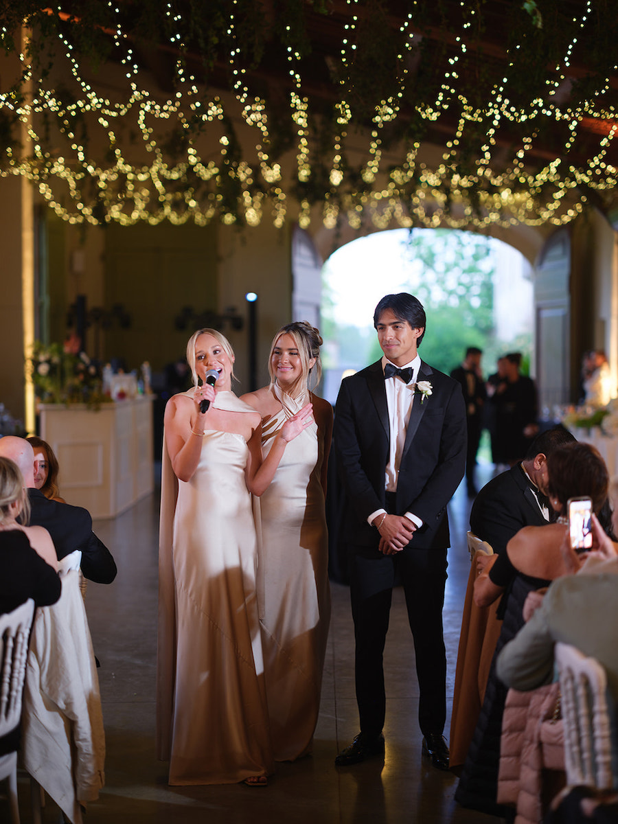 Two women in formal dresses and a man in a tuxedo stand together at a decorated indoor event, while guests seated at tables watch and take photos.
