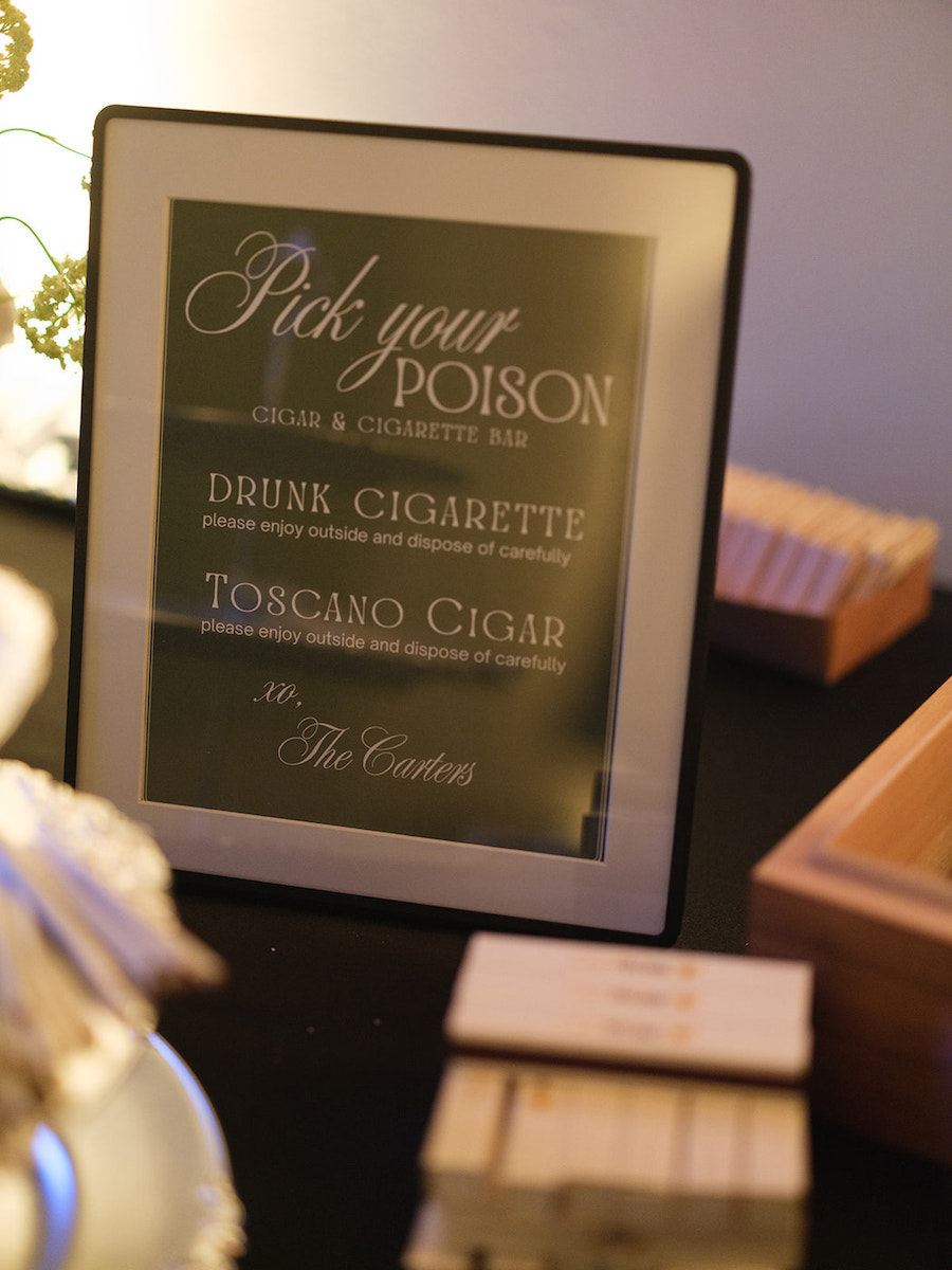 Framed sign reads “Pick your poison” with options for cigars and cigarettes on a table with boxes and matchsticks nearby.