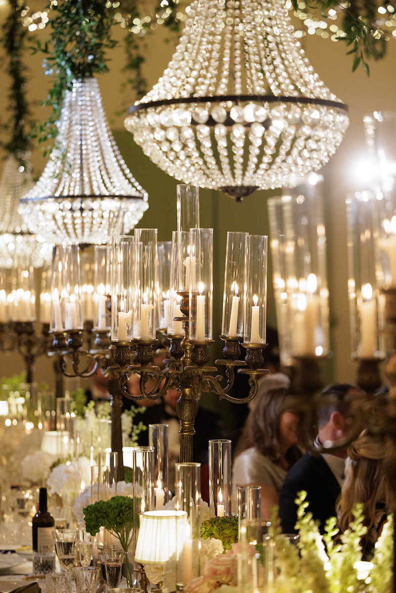Elegant chandeliers and candelabras with lit candles hang above a formal dinner table decorated with glassware, flowers, and greenery.