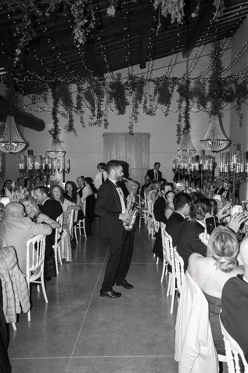 A saxophonist performs between long tables of seated guests at an indoor event decorated with hanging greenery and chandeliers.