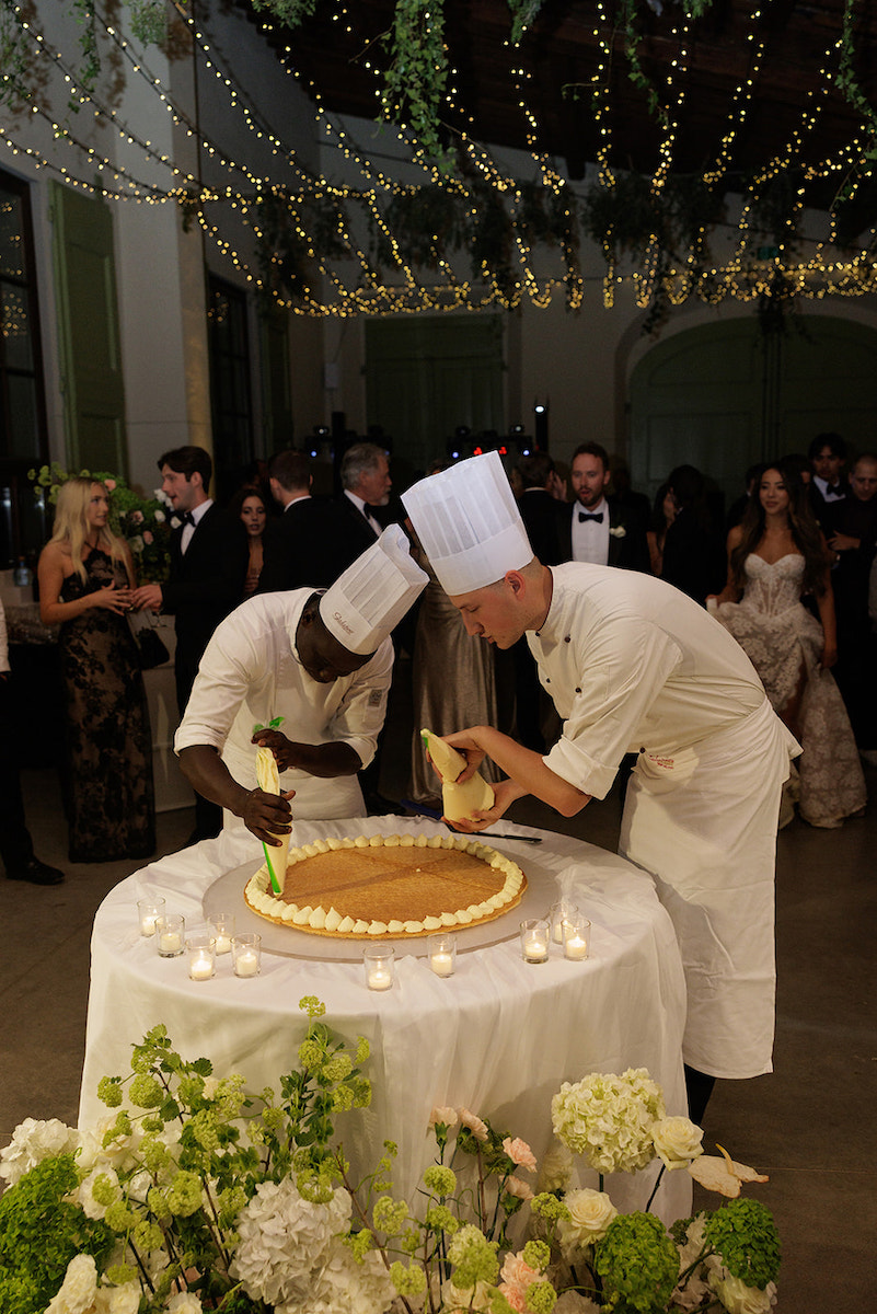 Two chefs in white uniforms and hats decorate a large round cake on a table, surrounded by flowers and candles, with guests watching in the background.