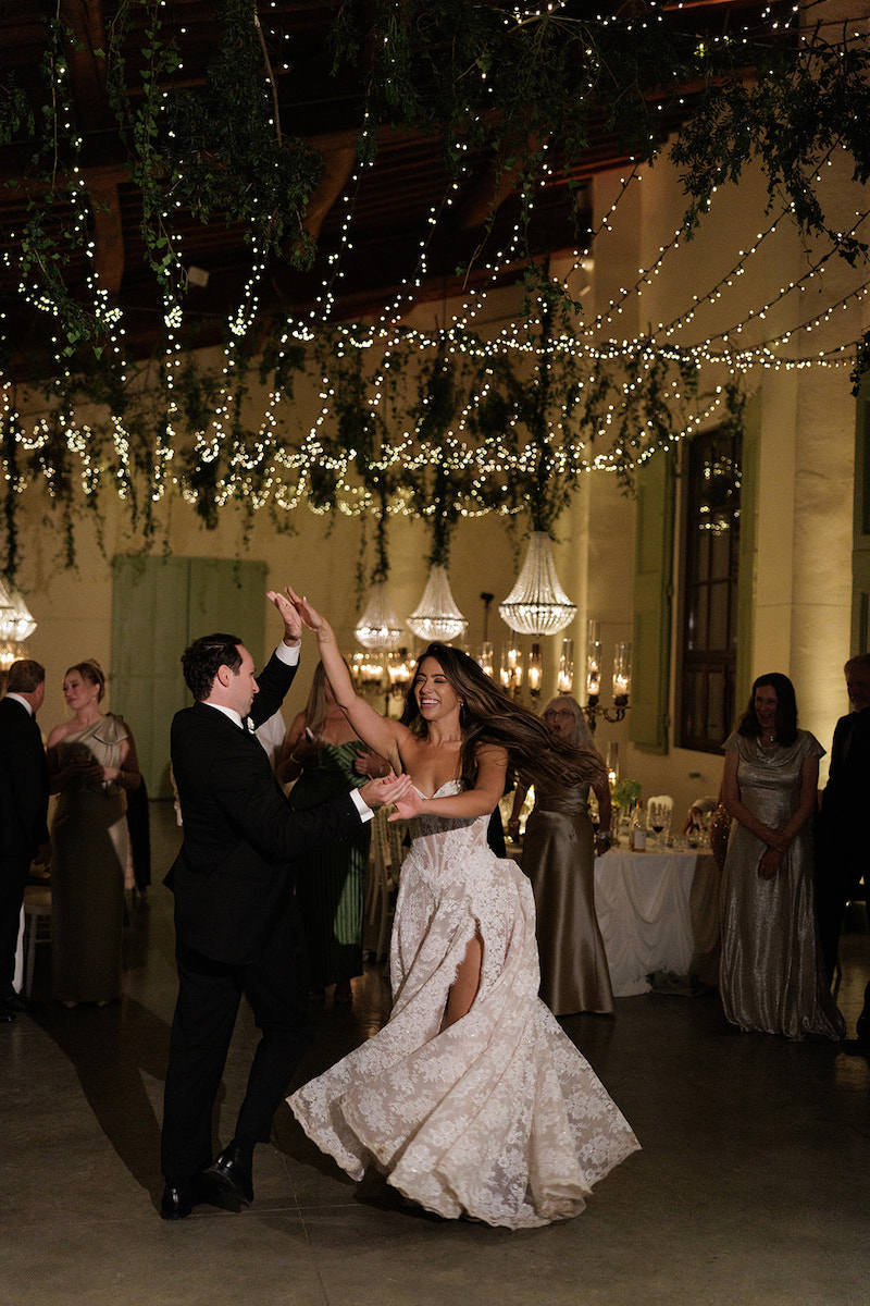 A bride and groom dance together at their wedding reception under hanging lights and greenery, with guests watching in the background.