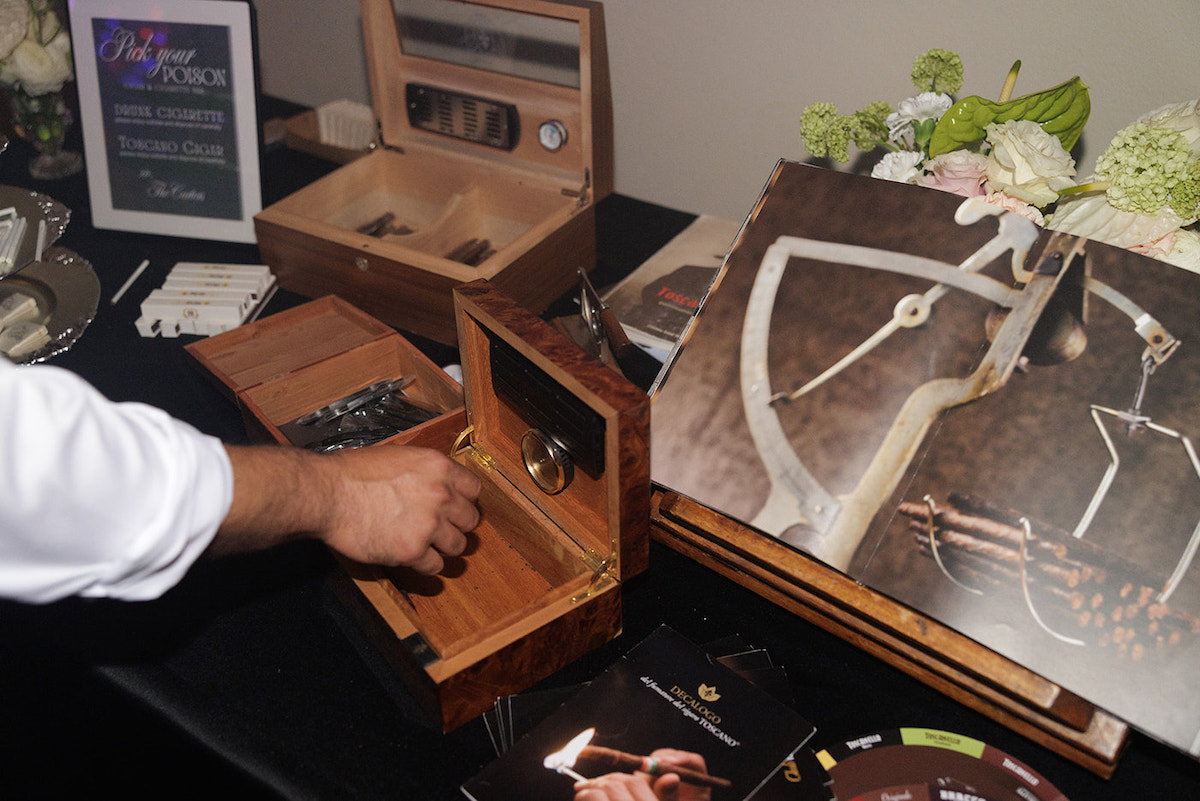 A person’s hand reaches into a wooden humidor box on a table with cigars, cigar accessories, an open book, and a floral arrangement in the background.