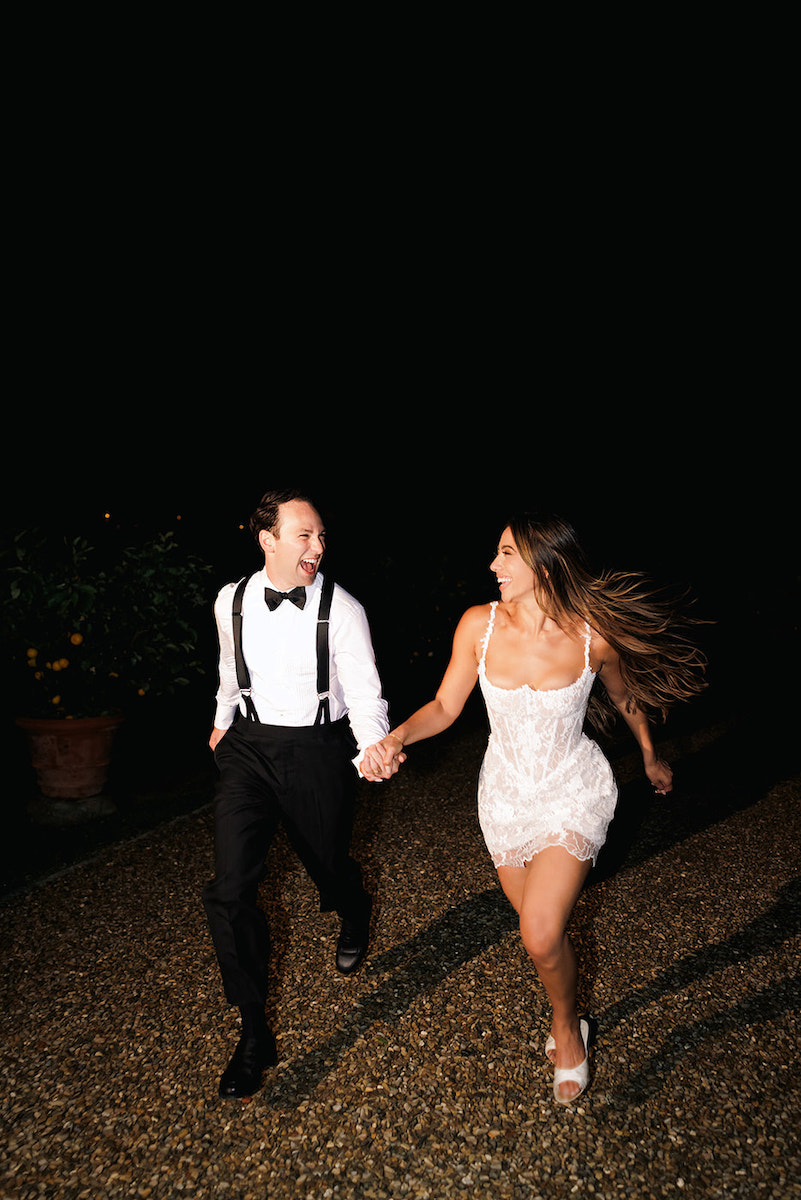 A man in formalwear and a woman in a short white dress run hand in hand at night on a gravel path, both smiling and looking at each other.
