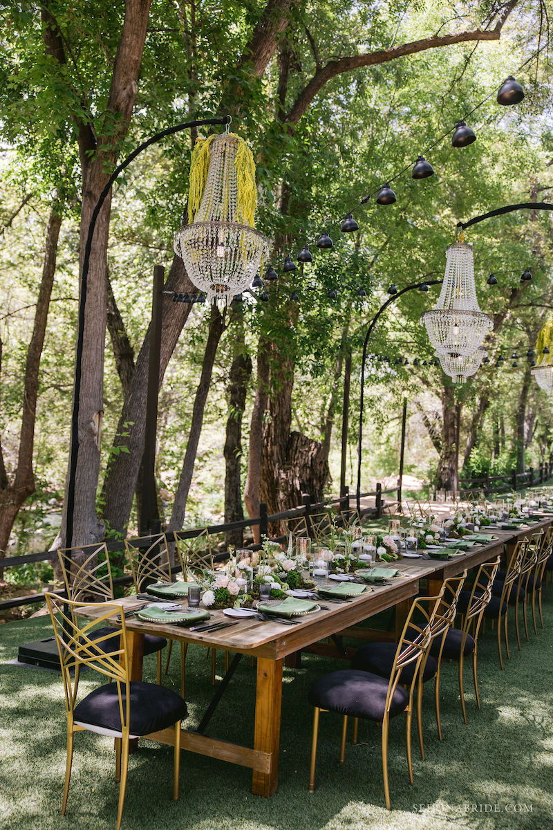 Long wooden dining table set for a meal outdoors, surrounded by gold chairs, with chandeliers and string lights hanging above under tall trees.