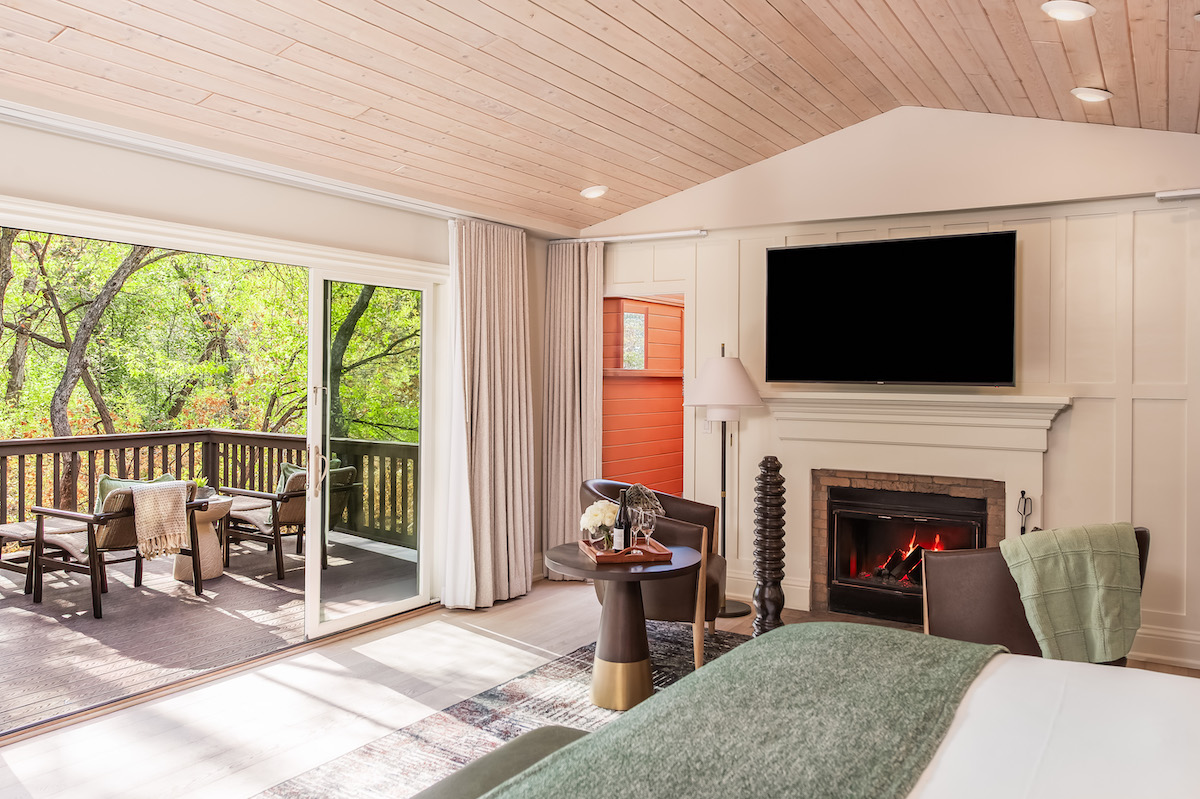 A hotel room with a bed, fireplace, wall-mounted TV, seating area, and sliding doors opening to a furnished balcony with a wooded view.