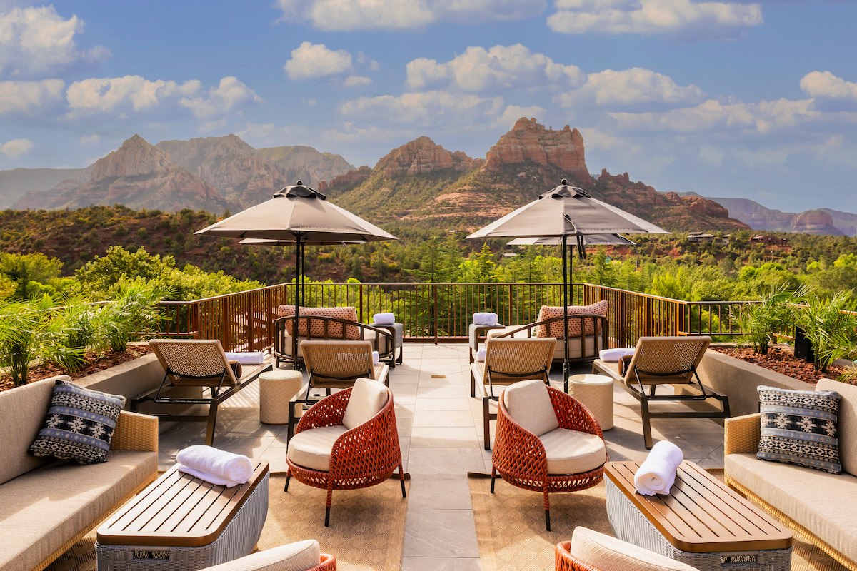 Outdoor lounge area with cushioned seating, tables, and umbrellas overlooks scenic rocky mountains under a blue sky with scattered clouds.