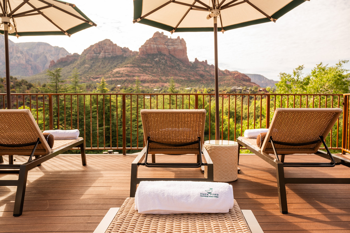 Four lounge chairs and umbrellas on a wooden deck overlook green trees and red rock mountains under a partly cloudy sky.