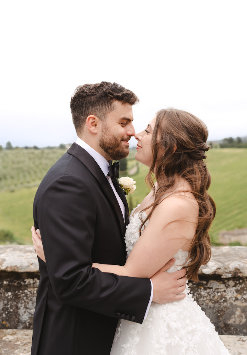 A bride and groom in formal wedding attire embrace outdoors near a stone wall, with green fields visible in the background.