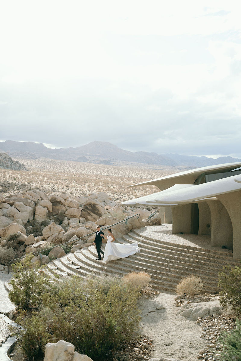 A bride and groom walk hand in hand down curved stone steps outside a modern building in a rocky desert landscape under a cloudy sky.
