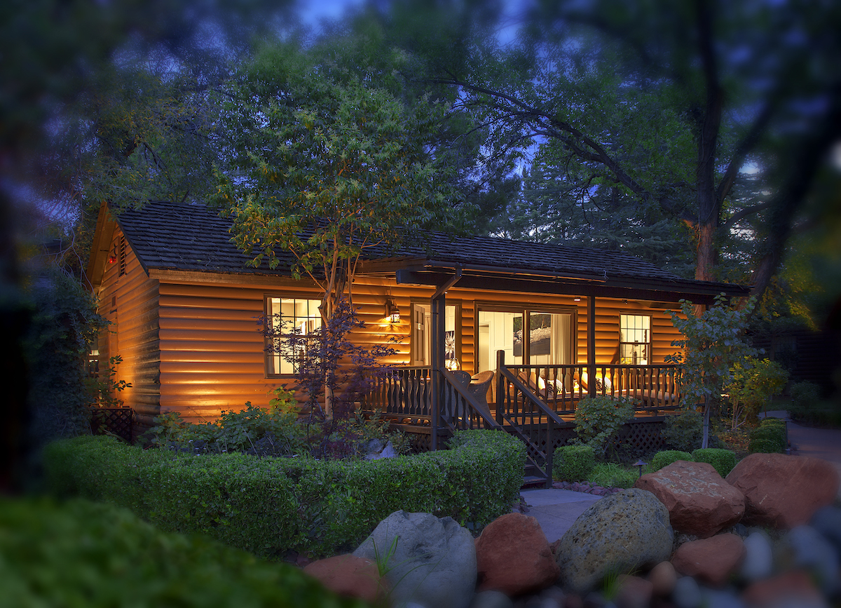 A small, warmly lit log cabin with a front porch is surrounded by shrubs, trees, and rocks at dusk.