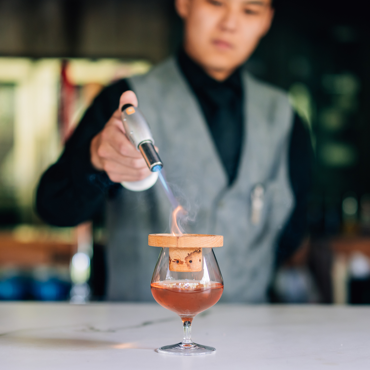 A bartender uses a torch to heat a wooden lid over a cocktail glass that contains a drink and a large ice cube.