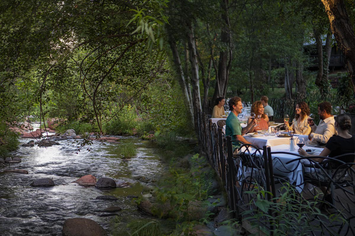 A group of people dine at a table outdoors in a lush, green forest setting next to a gently flowing stream.