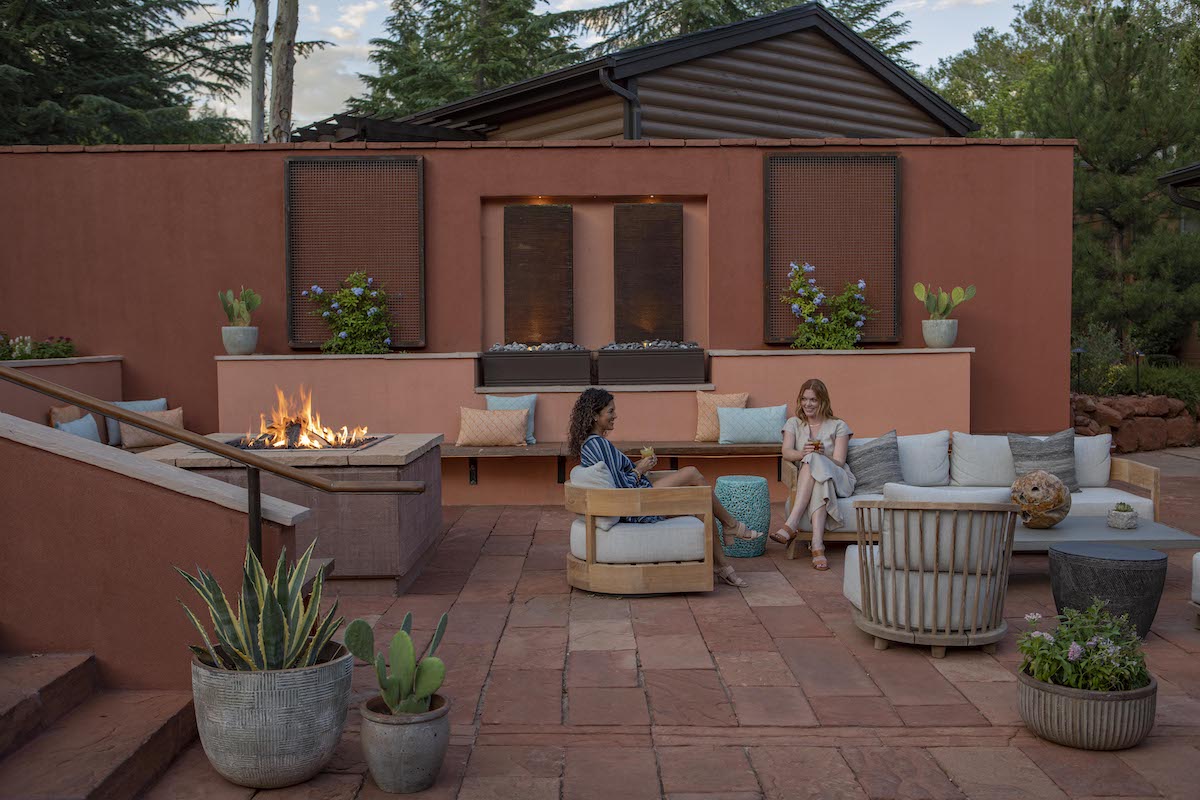 Two people sit and talk on outdoor patio furniture near a firepit, surrounded by potted plants, with a terracotta-colored wall and building in the background.