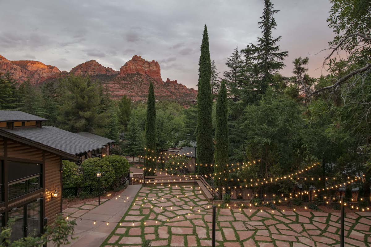 A courtyard with stone paving and string lights is surrounded by trees, with rocky mountains visible in the background under a cloudy sky at sunset.