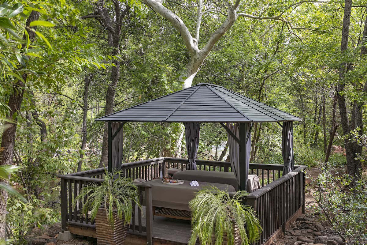 A small gazebo with a metal roof sits on a wooden deck in a forested area, surrounded by trees and potted plants.