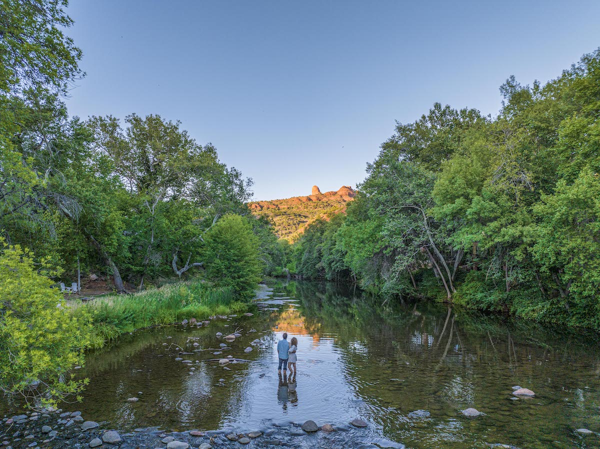 Two people stand in a shallow creek surrounded by dense green trees, with a hill and clear blue sky in the background.