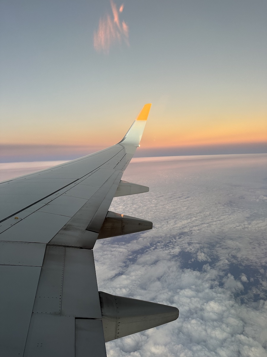View from airplane window showing the wing and winglets above a blanket of clouds during sunset, with an orange and pink sky in the background.