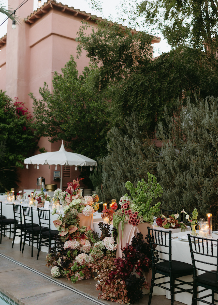 Long outdoor dining table with black chairs, floral arrangements, candles, and a white umbrella beside a pink stucco building, surrounded by greenery.