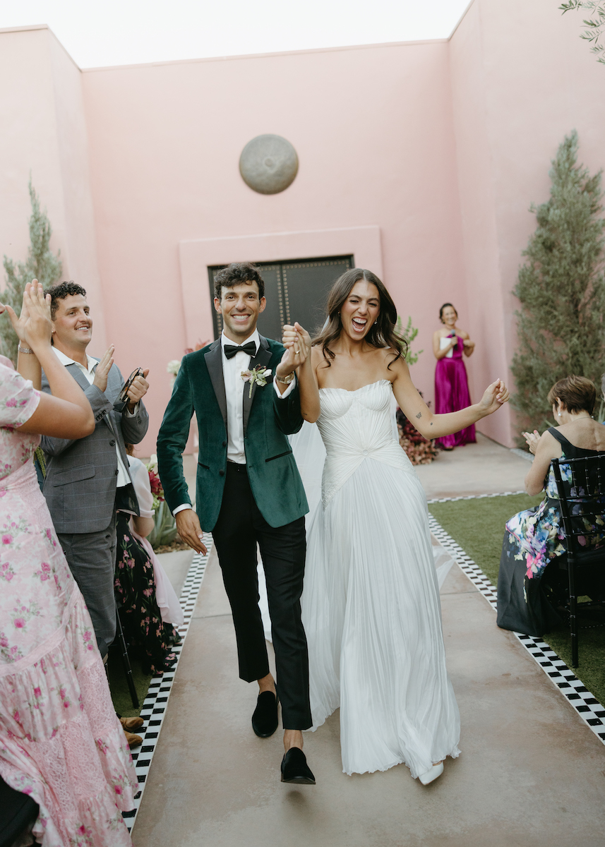 A bride and groom walk down an outdoor aisle, smiling and holding hands, while guests clap and celebrate their wedding.