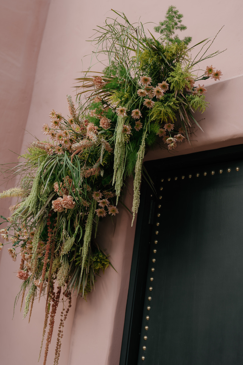 Greenery and dried flowers arranged above a dark door with metallic studs, set against a pale pink wall.