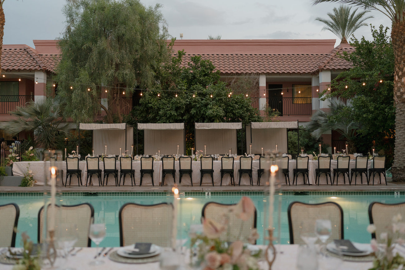 A long dining table with chairs is set up beside a swimming pool, with candles and place settings, in front of a building with palm trees and greenery.