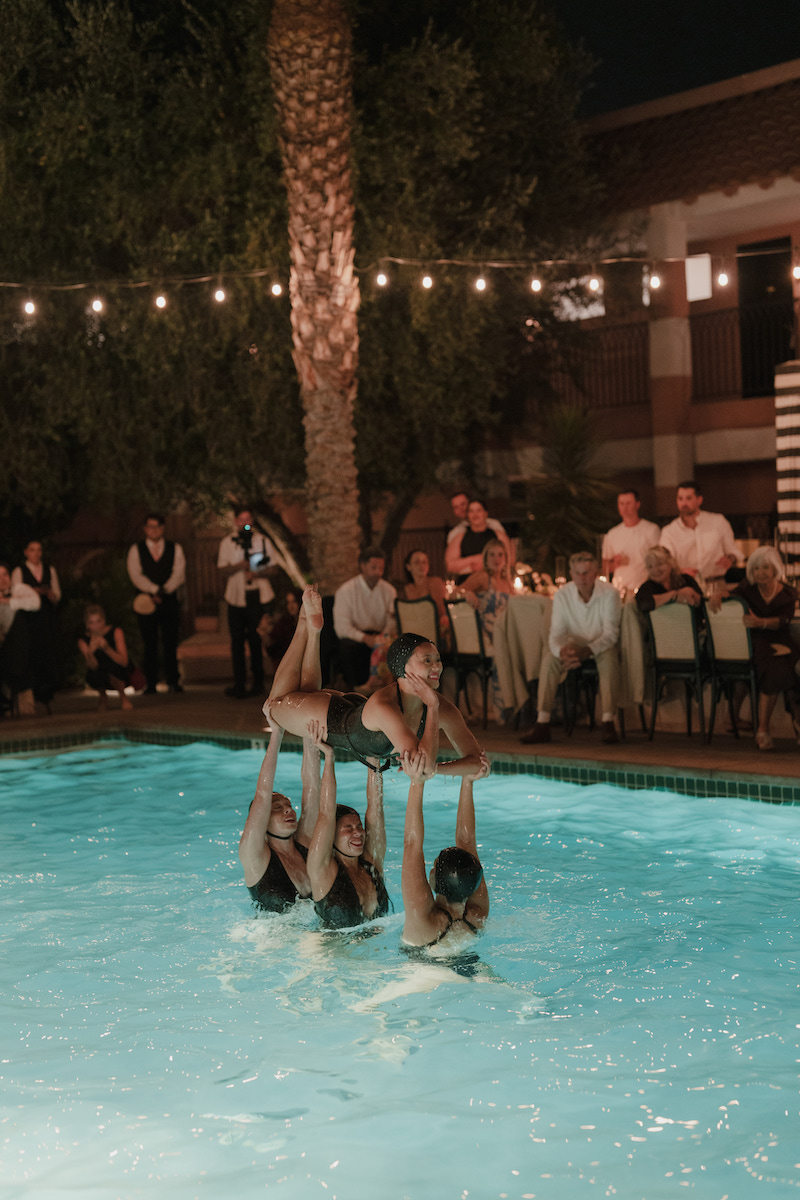 Three synchronized swimmers perform a lift in a pool at night, with an audience of formally dressed people watching from the poolside.