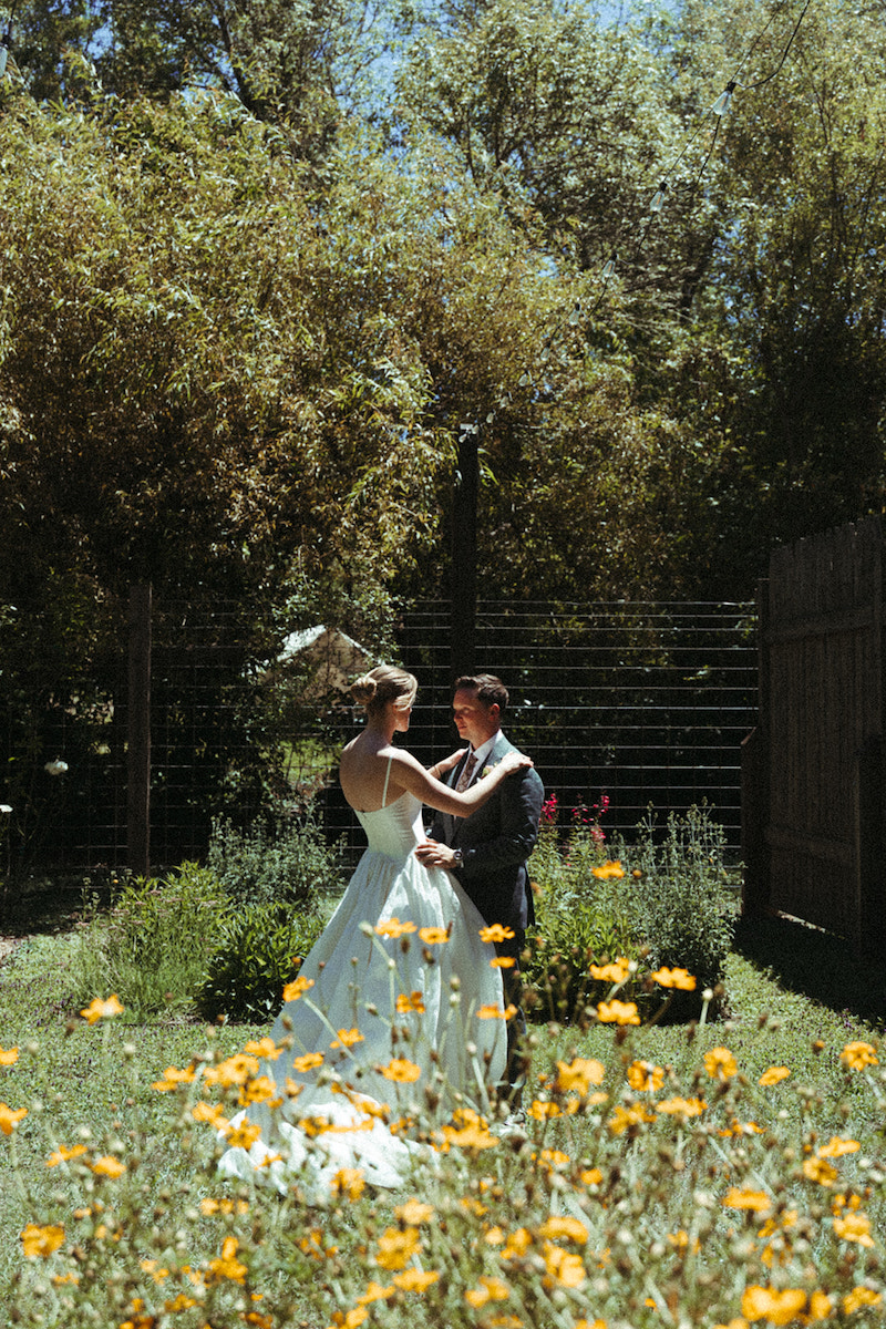 A bride and groom stand together in a sunlit garden surrounded by yellow flowers and greenery.