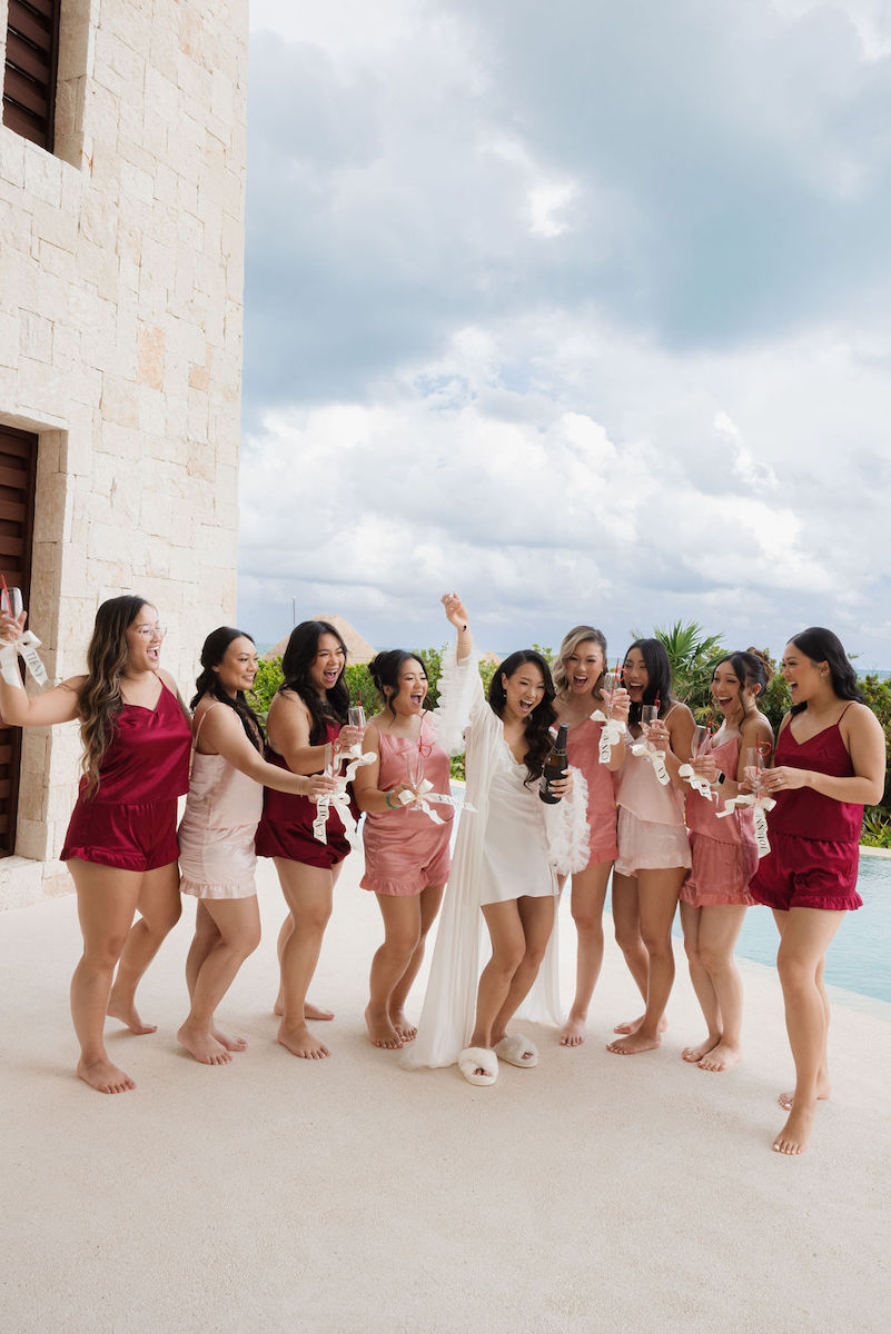 A group of women in pink and red outfits stands barefoot by a pool, laughing and holding drinks, while one woman in white celebrates in the center.