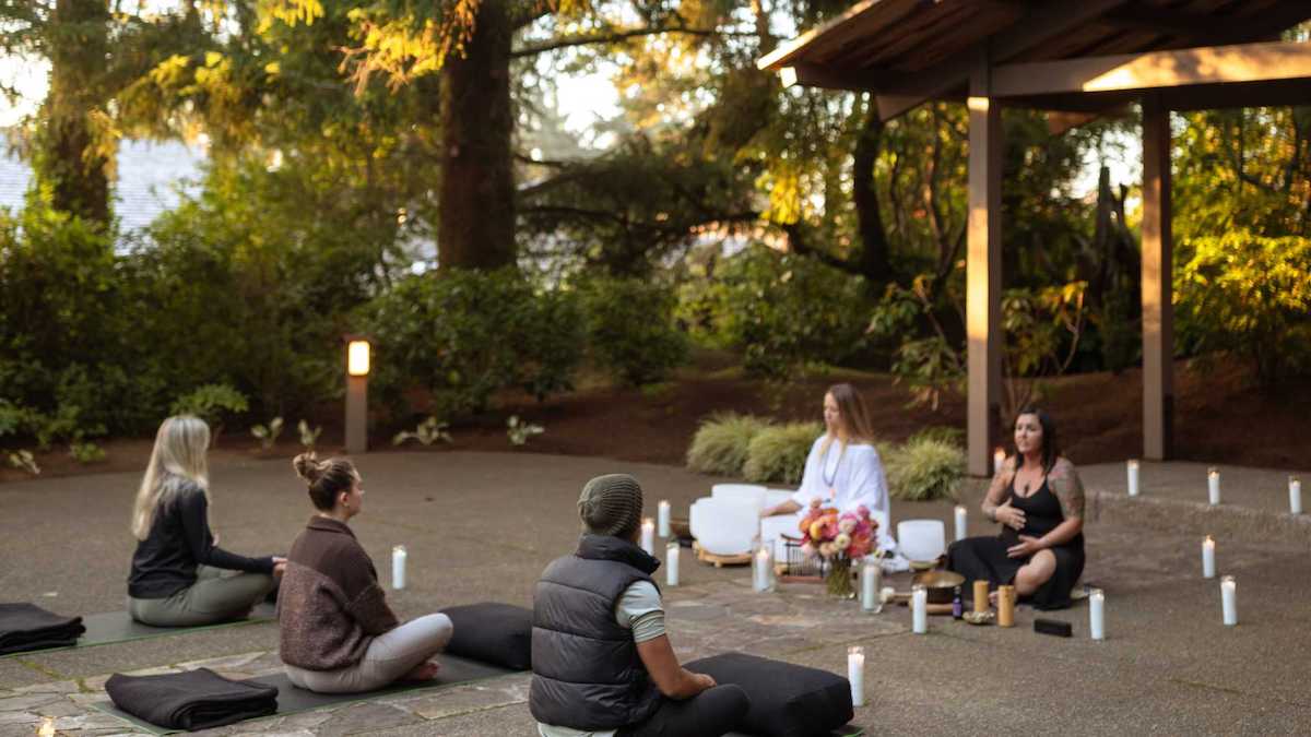 Five people sit on cushions in a circle outdoors, surrounded by trees and candles, participating in a meditation or group session near a wooden shelter.