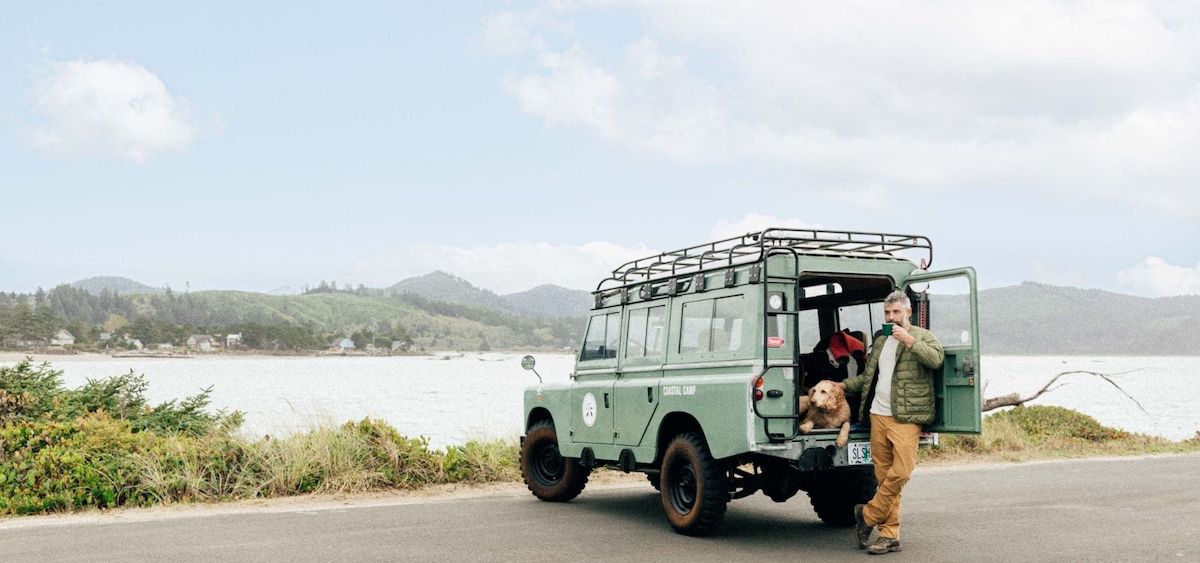 Person standing by an open vintage SUV with a dog inside, parked near a lake or sea with mountains and cloudy sky in the background.