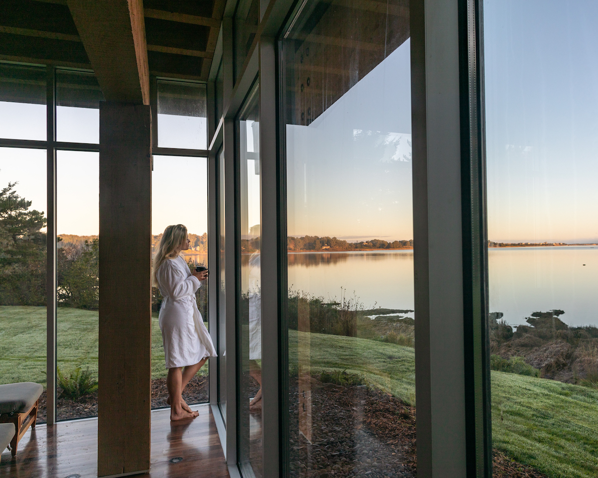 A person in a white robe stands by large windows, looking out at a calm lake and grassy landscape during sunrise or sunset.