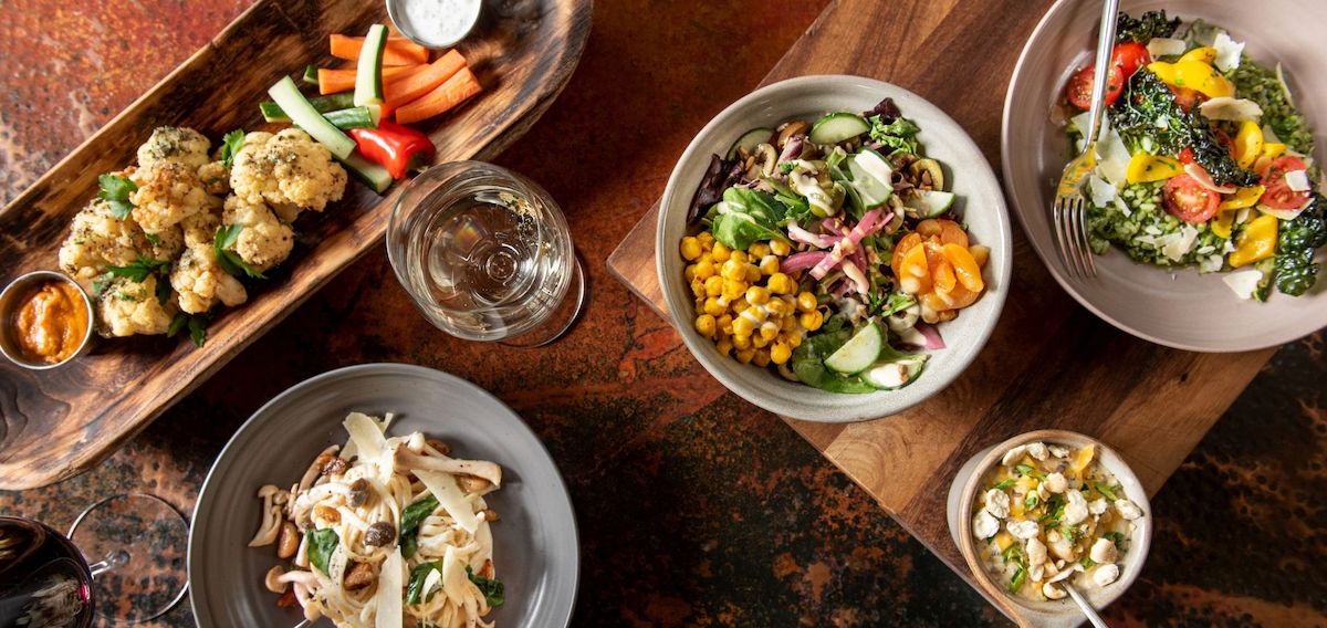Assorted dishes on a table, including salads with vegetables, a pasta dish, roasted cauliflower, dip, and a small bowl of salad, with glasses of water and wine.