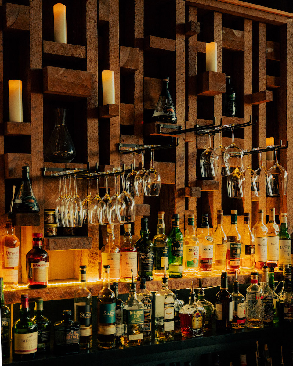 Bar shelves with bottles of liquor, hanging wine glasses, and candles on a wooden geometric background.