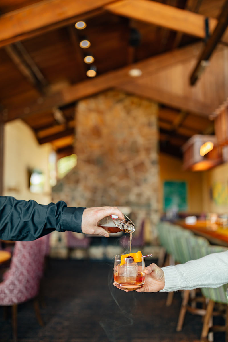A person pours a drink from a jigger into a glass with ice and an orange slice, inside a rustic bar with wooden beams and a stone fireplace.