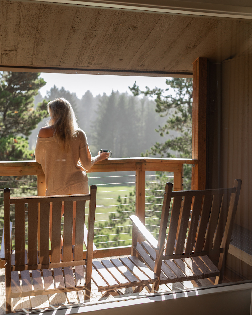 A person stands on a wooden balcony holding a mug, looking out at trees and a sunlit forest clearing. Two empty wooden chairs are in the foreground.
