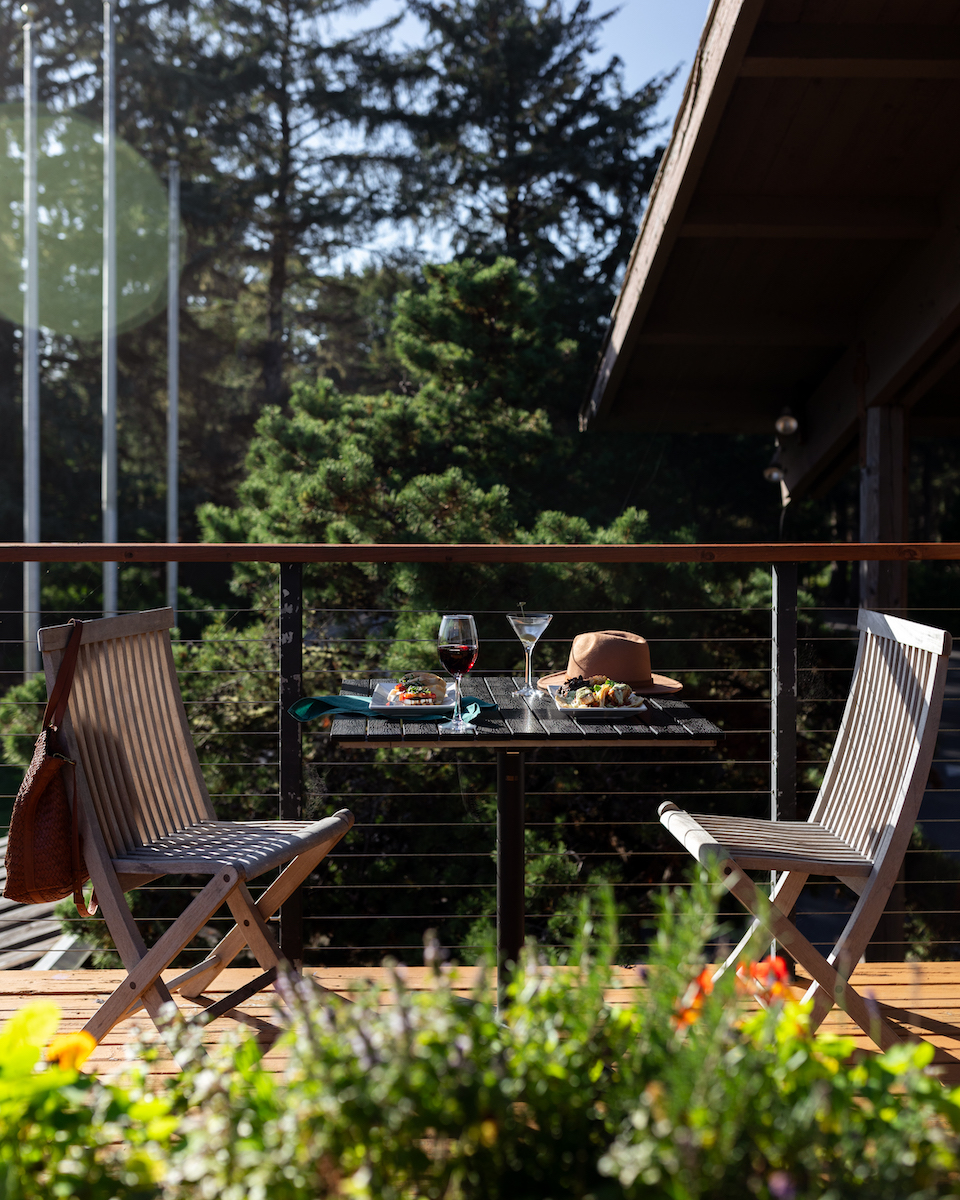 Two wooden chairs and a table with drinks and food are set on a balcony overlooking trees, with sunlight streaming in.