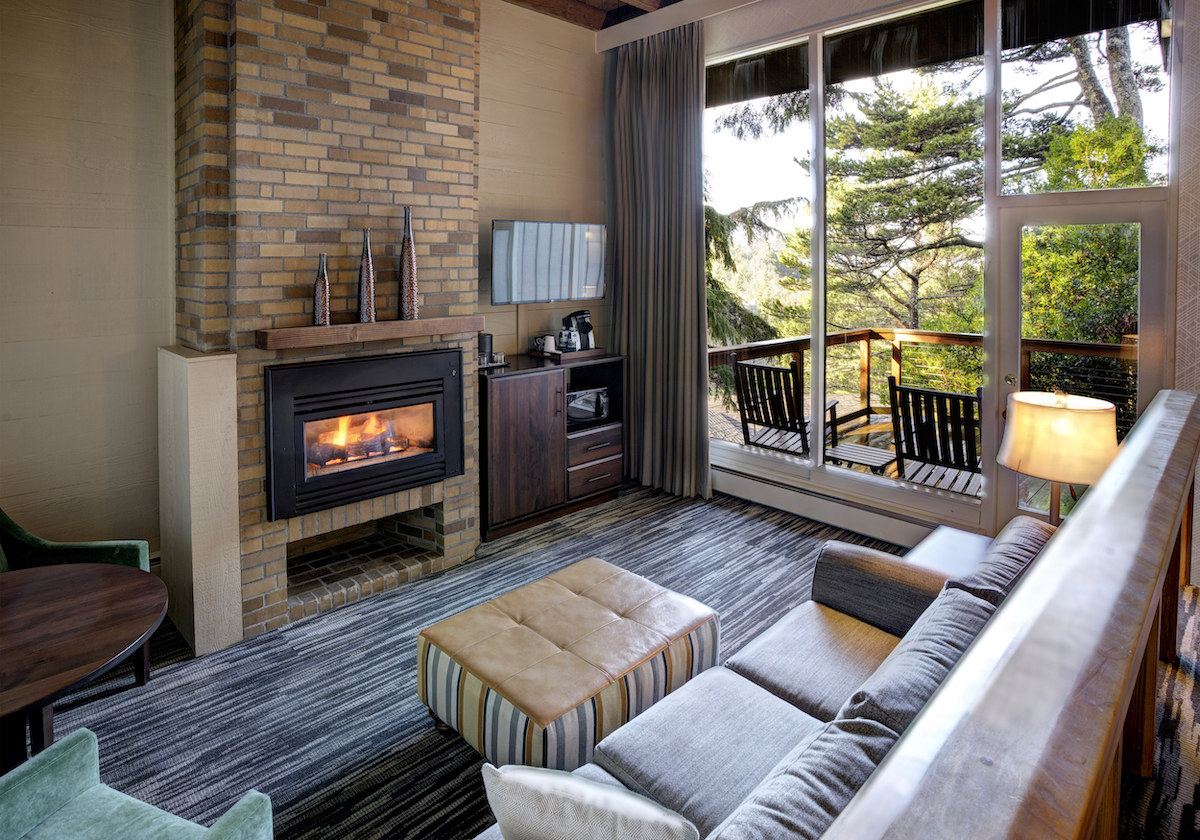 Modern living room with a fireplace, couch, striped ottoman, TV, and large windows overlooking a balcony with chairs and trees outside.
