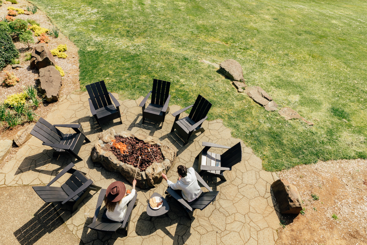 Two people sit by a fire pit surrounded by empty Adirondack chairs on a stone patio, with a green lawn and landscaping nearby.