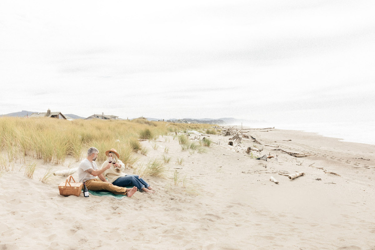 Two people sit on a sandy beach with a dog near grassy dunes, enjoying a picnic under a cloudy sky.