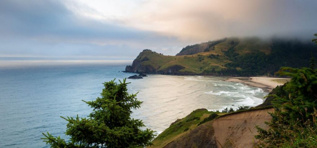 A coastal landscape with a sandy beach, ocean waves, rocky cliffs, and a misty hill under a cloudy sky.