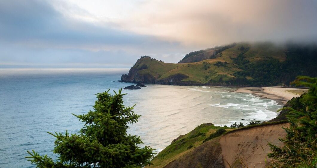 A coastal landscape with a sandy beach, ocean waves, rocky cliffs, and a misty hill under a cloudy sky.