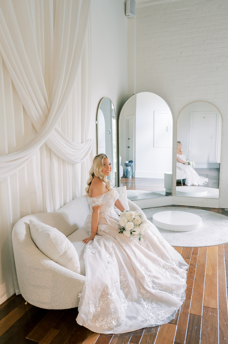 A bride in a white gown sits on a curved white sofa, holding a bouquet, with three tall mirrors and draped white fabric in the background.