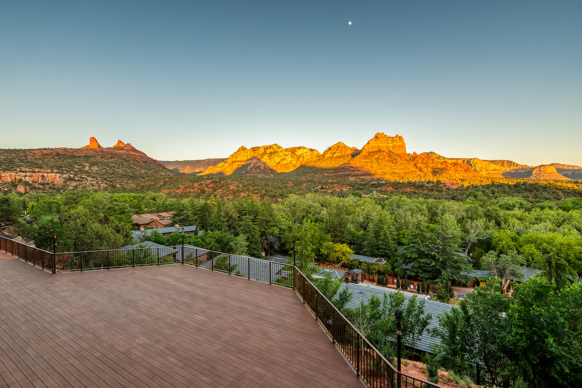 Wooden deck overlooking green trees and a valley with red rock mountains in the distance, lit by the setting sun under a clear sky with a visible moon.