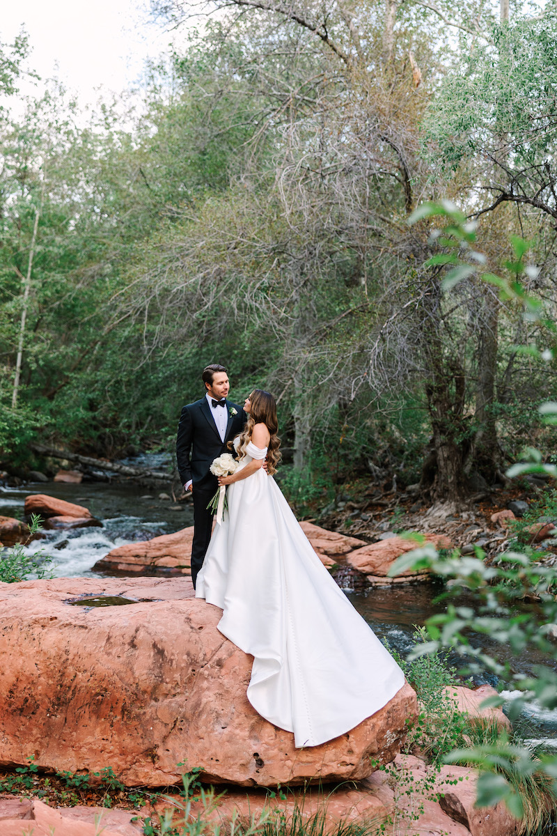 Bride and groom stand together on a large red rock beside a stream, surrounded by trees and greenery.